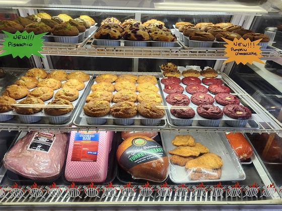 The bakery case stacked with trays of jumbo muffins and cookies, with handwritten signs reading New Pistachio and New Pumpkin Cheesecake