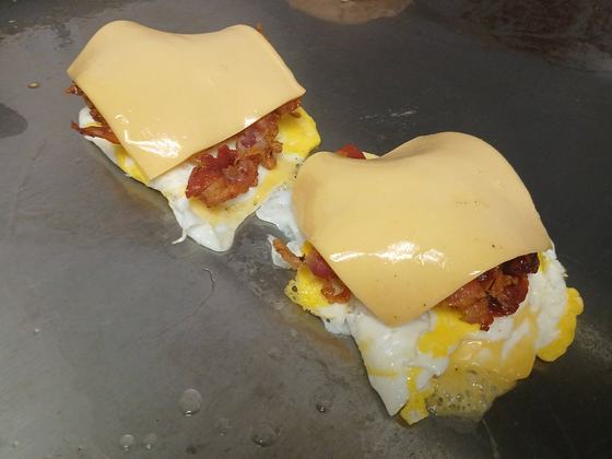 Two breakfast sandwiches on the griddle: fried eggs topped with bacon and slices of American cheese melting over them