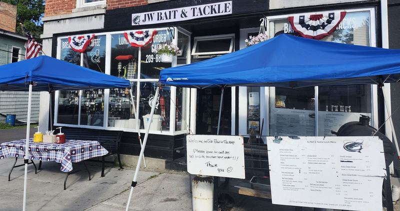 The JW Bait & Tackle storefront on a summer morning, flag bunting in the windows and two blue canopies over the sidewalk tables